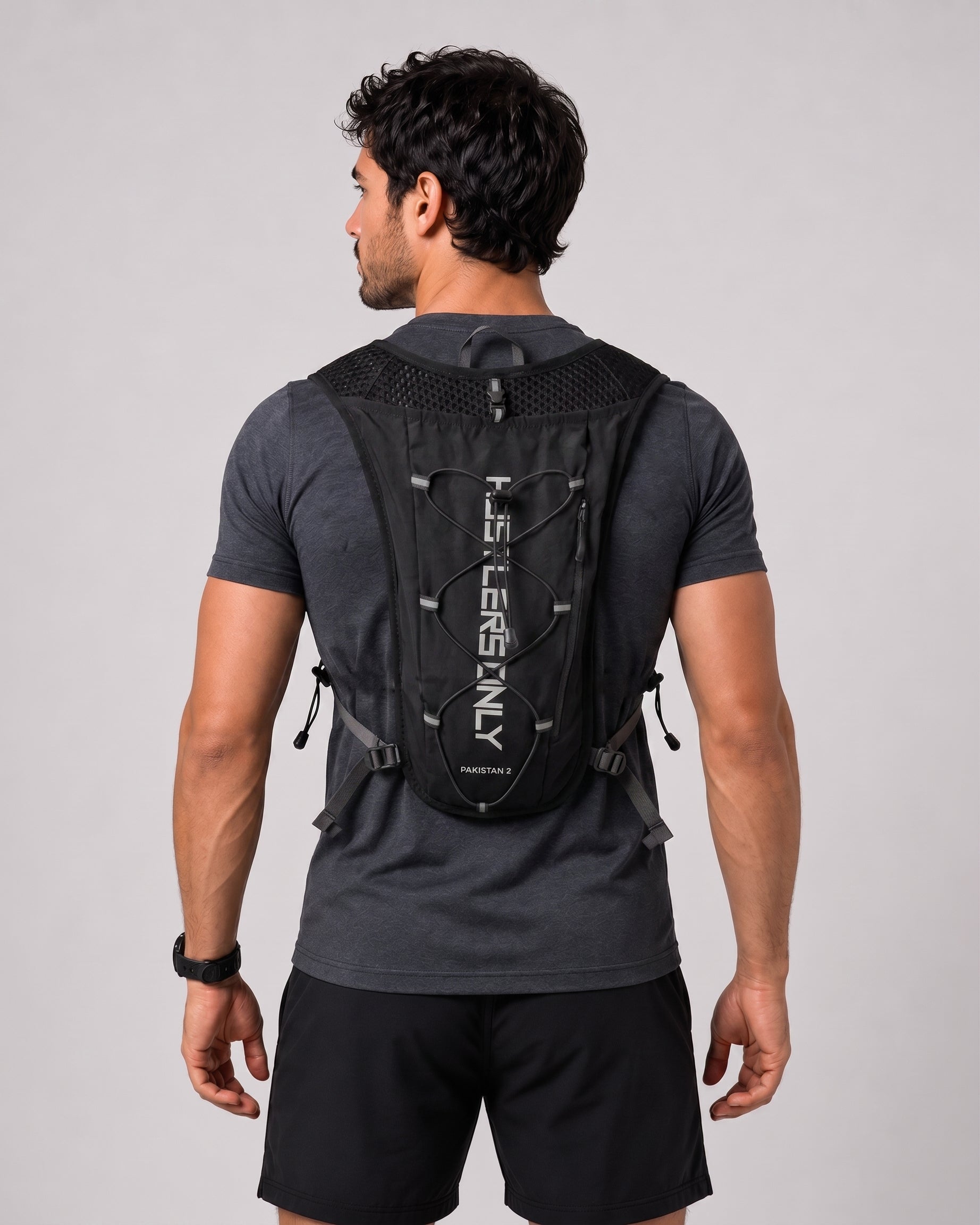 A man viewed from behind wearing a black TheroRun backpacking system with white vertical branding on the back, over a dark gray t-shirt and black shorts, against a light gray studio background.