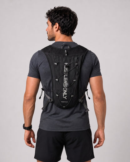 A man viewed from behind wearing a black TheroRun backpacking system with white vertical branding on the back, over a dark gray t-shirt and black shorts, against a light gray studio background.